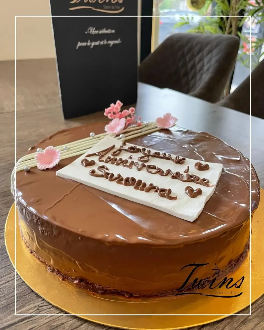 Un gâteau d'anniversaire au chocolat glacé, orné d'une plaque en chocolat blanc et de fleurs séchées, sur un plateau doré.