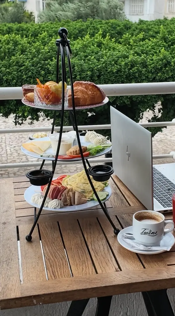 Un présentoir à trois étages en fer forgé sur une table en bois du balcon, à côté d'un café servi dans une tasse Twins.