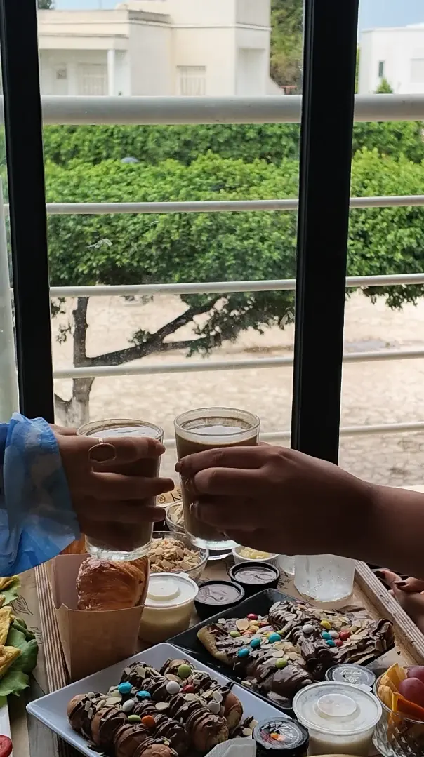 Deux mains trinquent avec leurs cafés au-dessus d'un brunch de crêpes au chocolat, devant la baie vitrée.